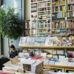 Young female client in casual wear chatting on cellphone while sitting at table with cup of hot drink in cozy bookstore in afternoon, tags: literarische neuerscheinungen - pexel