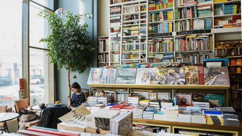 Young female client in casual wear chatting on cellphone while sitting at table with cup of hot drink in cozy bookstore in afternoon, tags: literarische neuerscheinungen - pexel